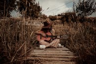 一位戴着帽子的女子在乡村草木间弹奏原声吉他，散发出一种宁静质朴的氛围