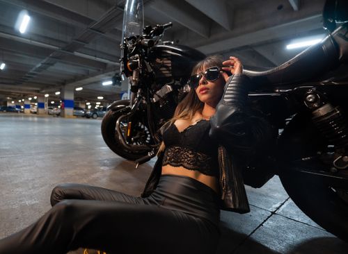 Free A fashionable woman poses with a motorcycle in a dimly lit urban parking lot, exuding confidence and style. Stock Photo