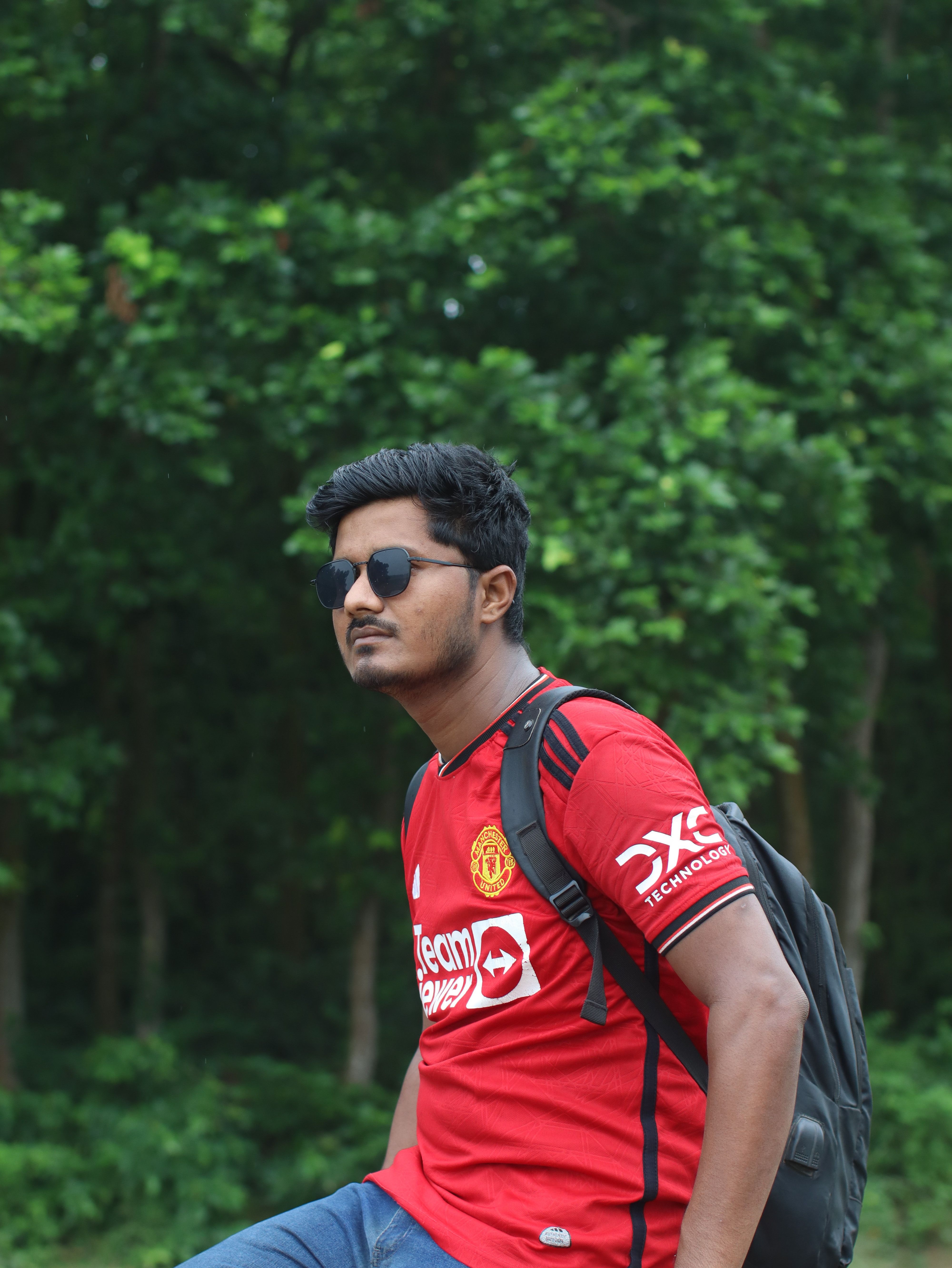 Young man wearing Manchester United jersey and sunglasses, posing outdoors with a backpack.