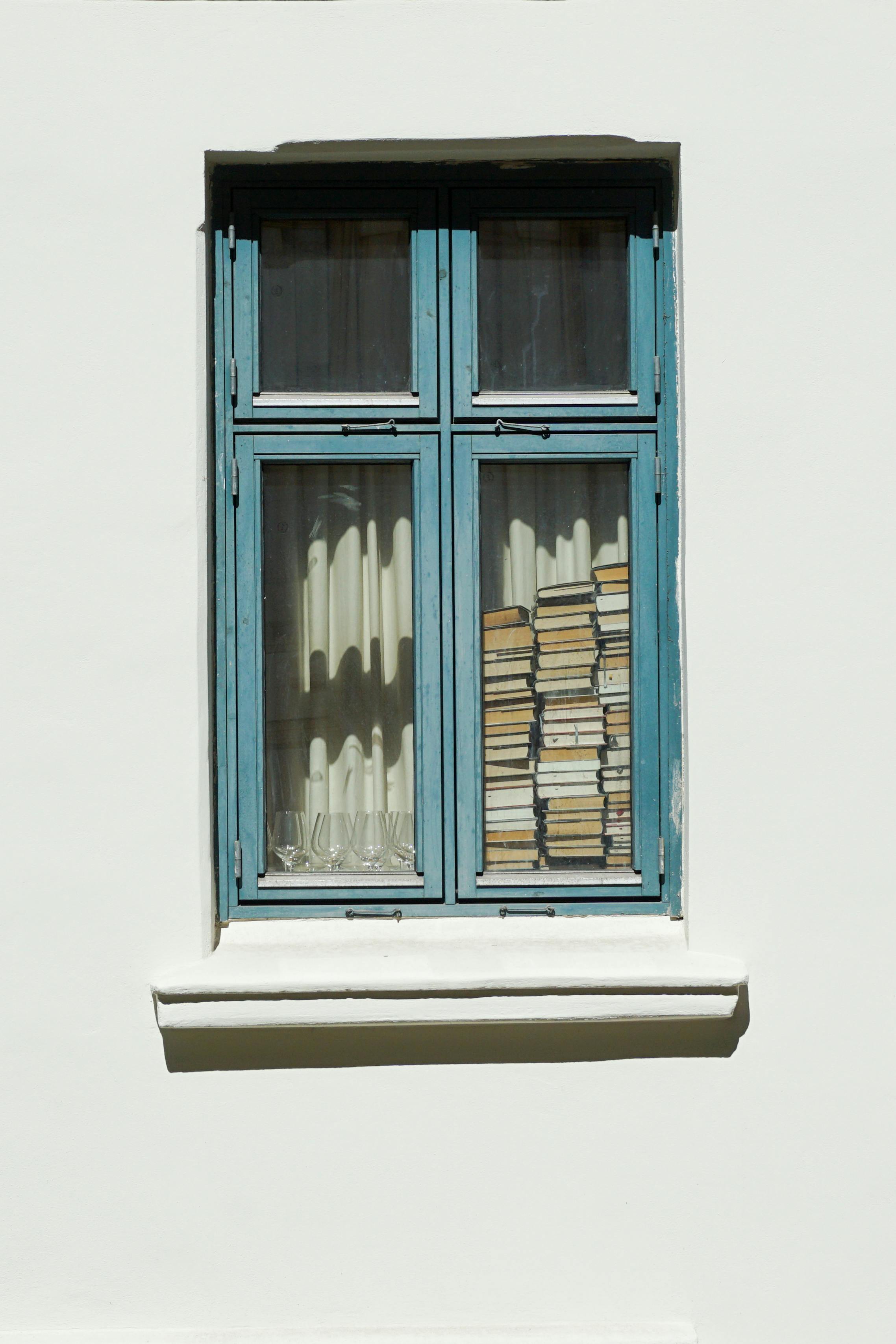 A stack of books seen through a vintage blue-framed window in Oslo, Norway, with soft sunlight casting shadows.