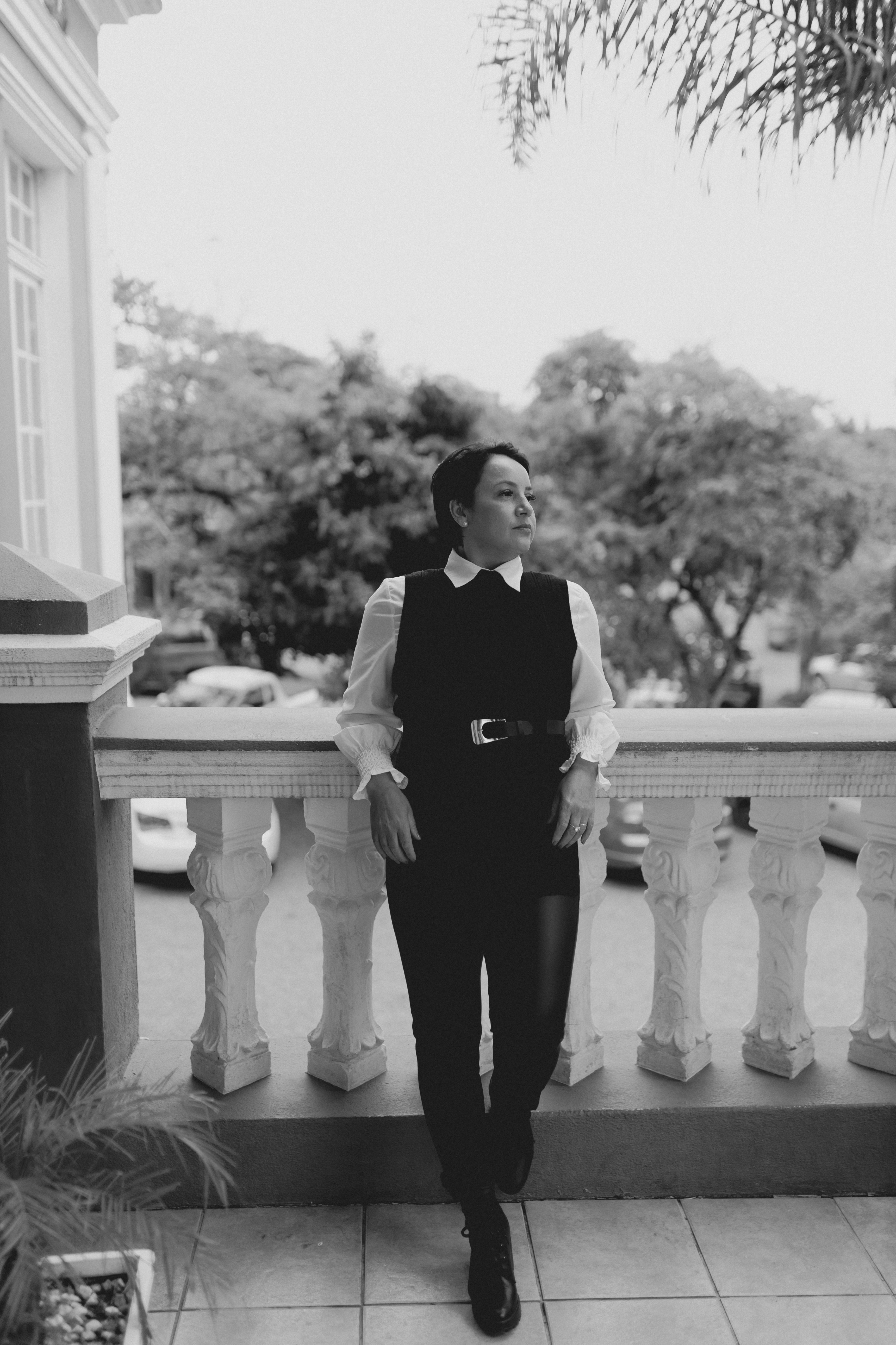Black and white photo of a woman in formal attire standing on a balcony, exuding elegance.