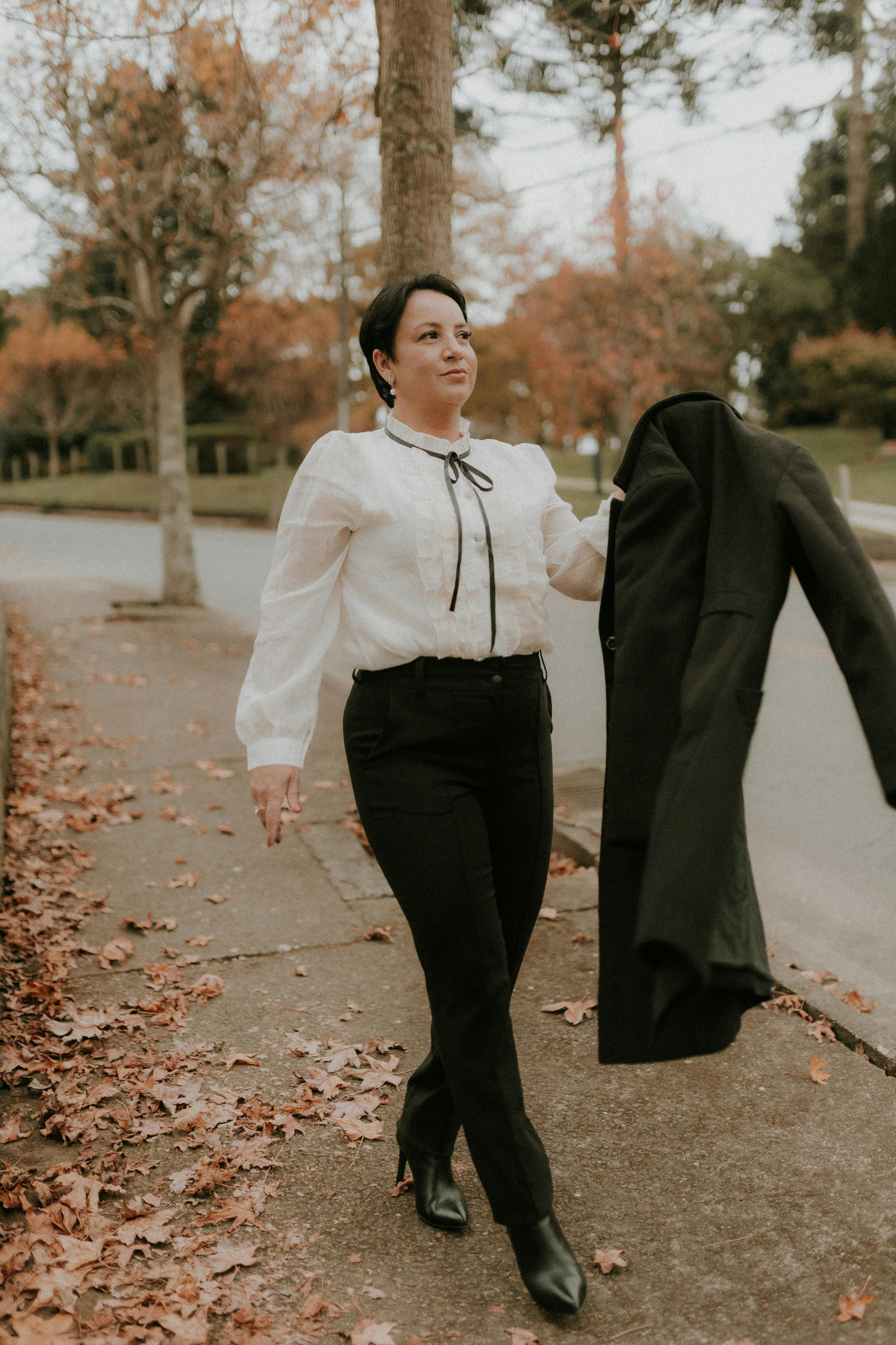 Elegant woman walking on an autumn street holding a coat, showcasing a stylish white blouse and heels.
