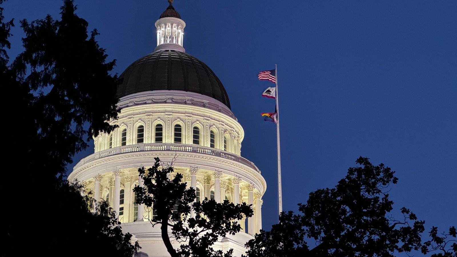 Illuminated California State Capitol dome against a clear night sky with fluttering flags.