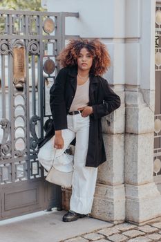Young woman with afro hair posing outdoors in stylish attire against a city fence.