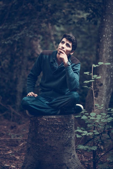 A man sits crossed-legged on a tree stump, with his hand on his chin, as thought thinking