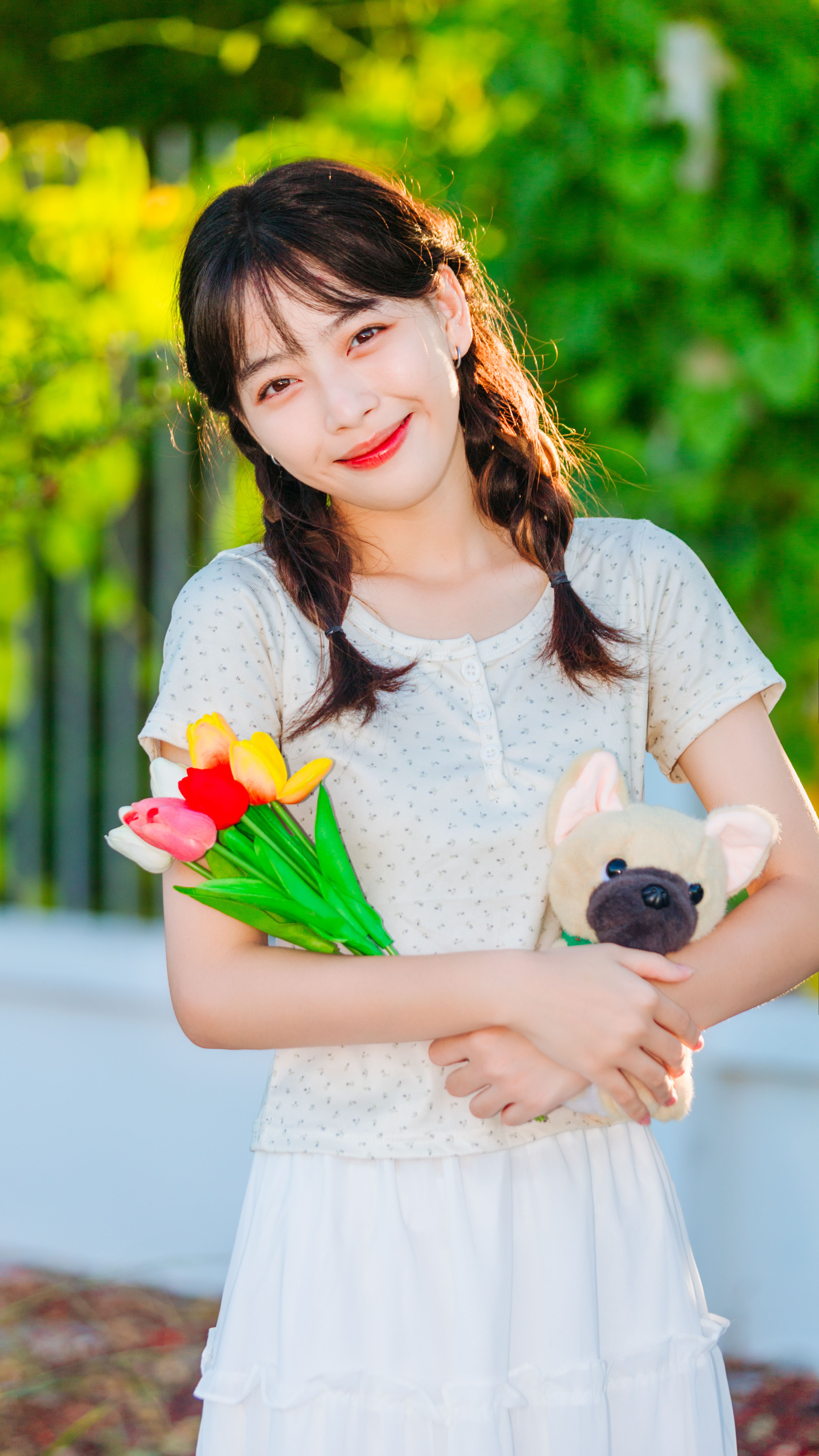 Smiling Asian woman holding colorful flowers and a stuffed toy outdoors.