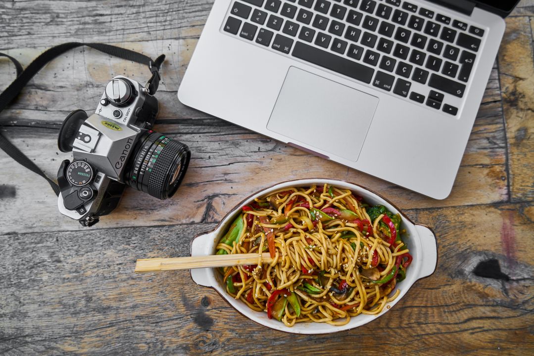 Recipe Developer Career in India: What Food Brands Actually Pay For 2 Overhead flat-lay of a DSLR camera, open laptop, and a bowl of noodles arranged together on a rustic wooden table