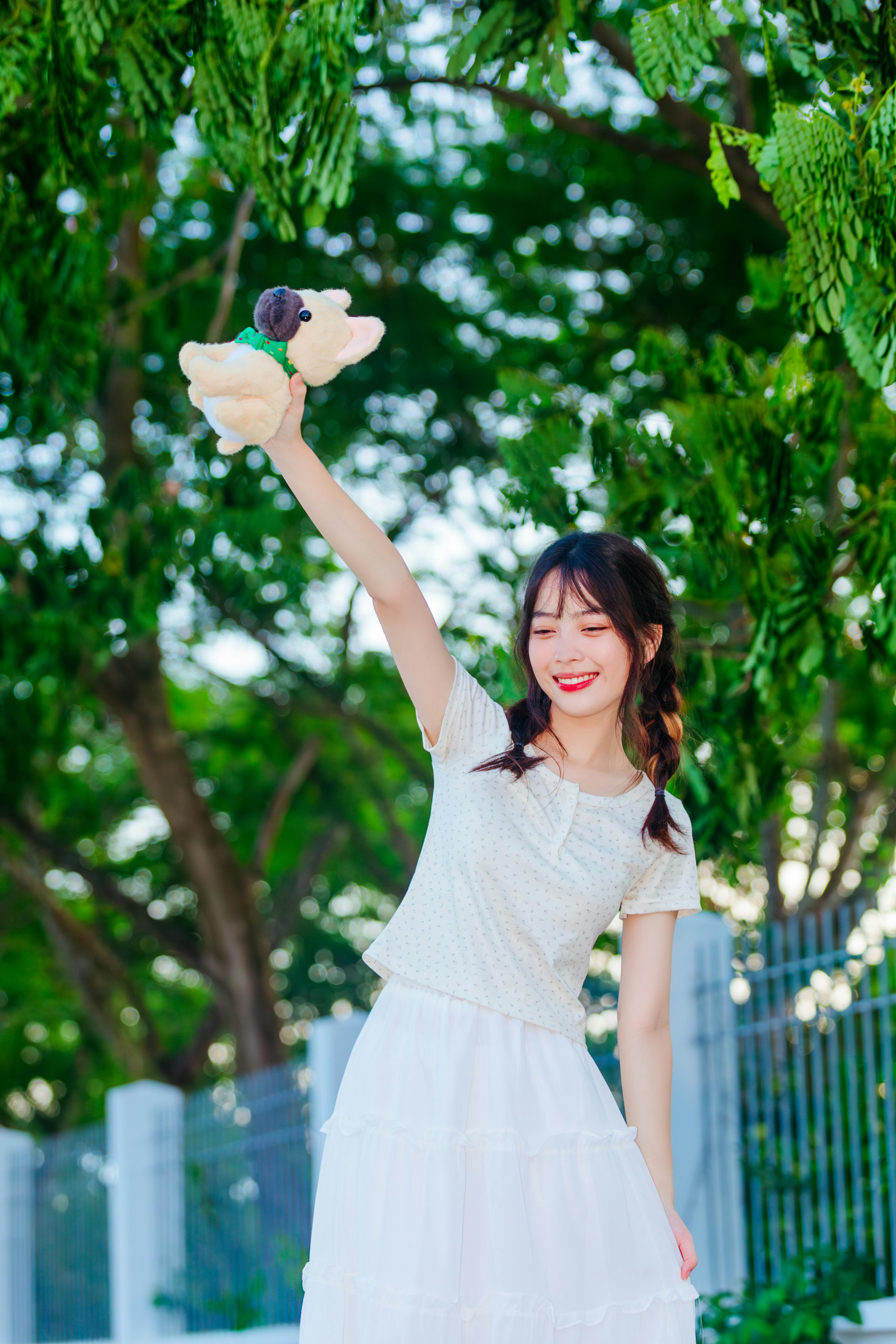 Asian woman standing outdoors with arm raised, holding a toy, surrounded by lush greenery.