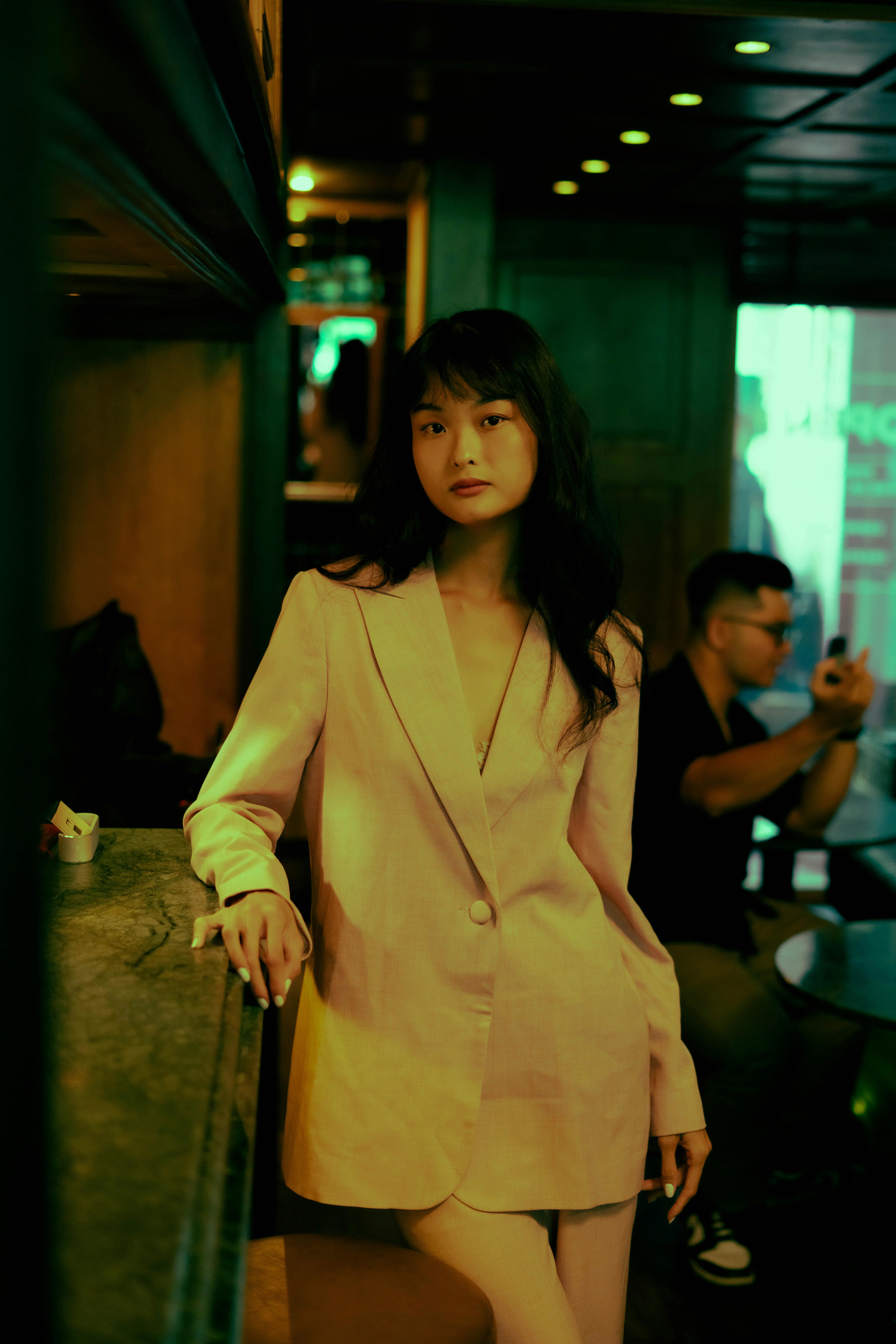 A serene woman in a fashionable jacket poses elegantly at a cozy bar.
