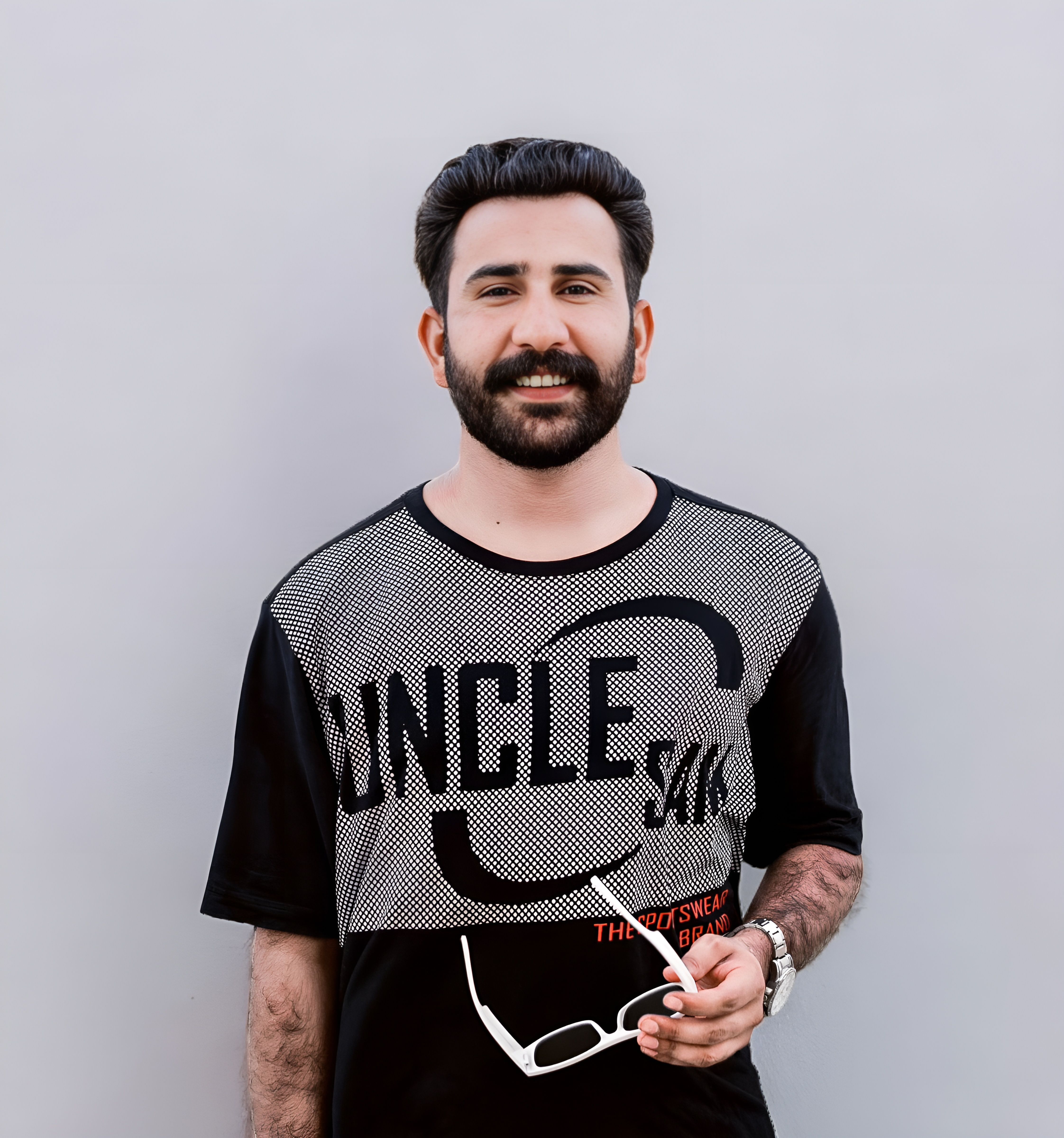 Stylish portrait of a man with a beard and sunglasses holding a T-shirt against a white background.