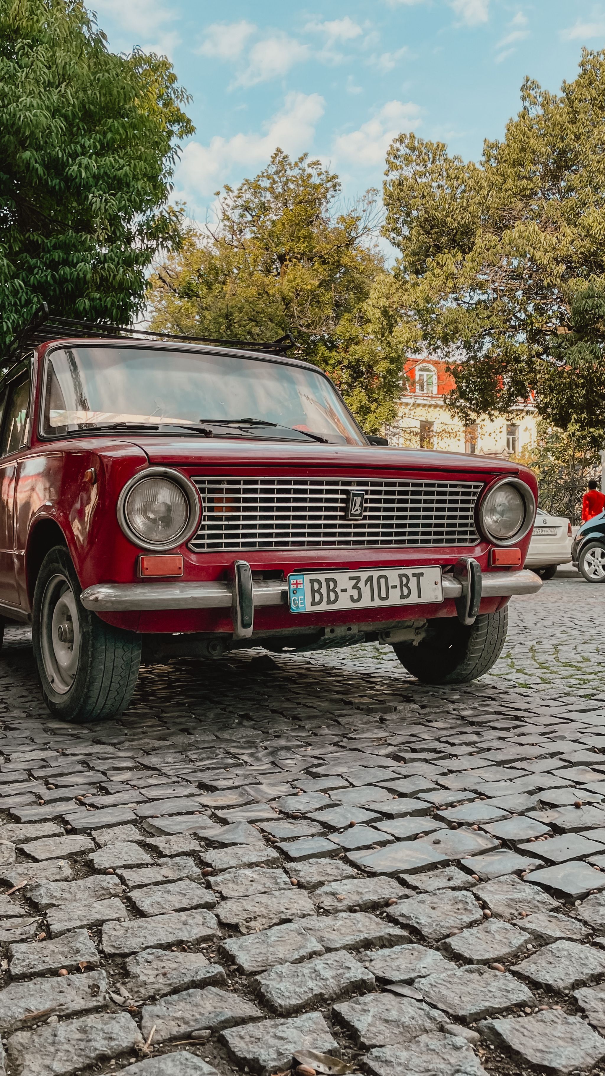 Red vintage car parked on cobblestone street with trees in an urban setting.