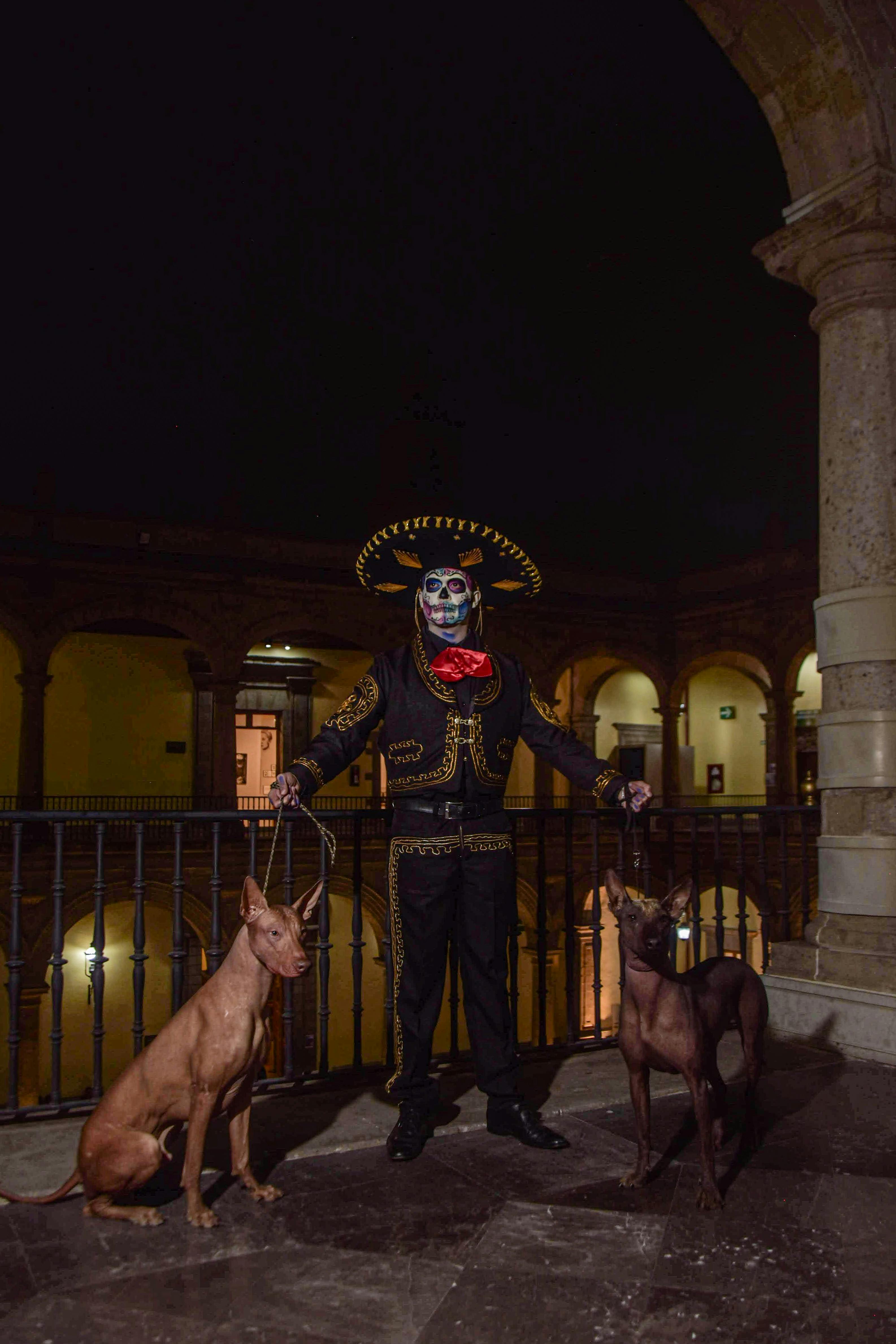 A man in Dia de Muertos attire with sugar skull face paint and dogs, in an urban setting at night.