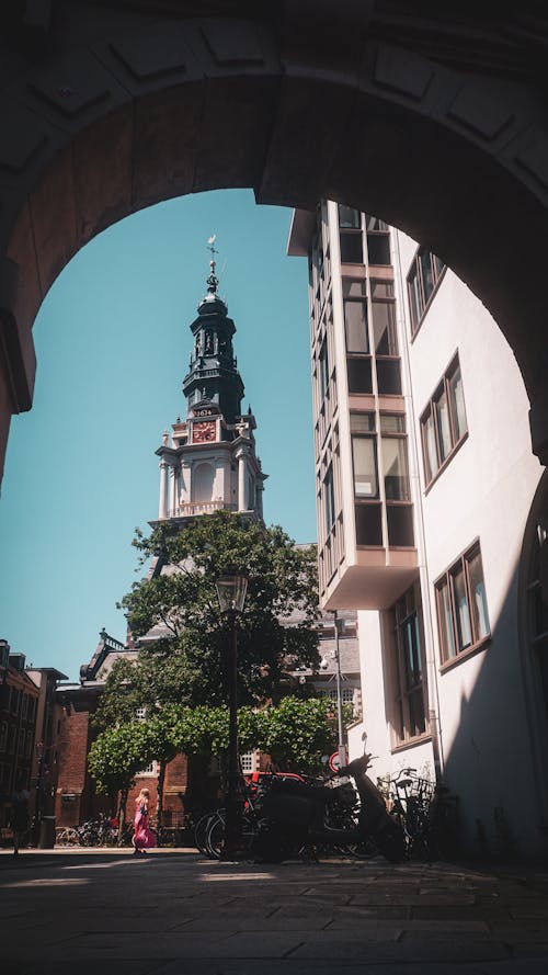 Free View of a historic church tower in Amsterdam framed by an archway, capturing urban life. Stock Photo