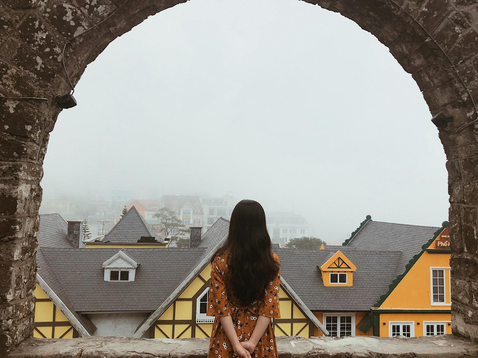 A woman in a floral dress gazes at a misty townscape through an ancient stone archway, creating a serene scene.