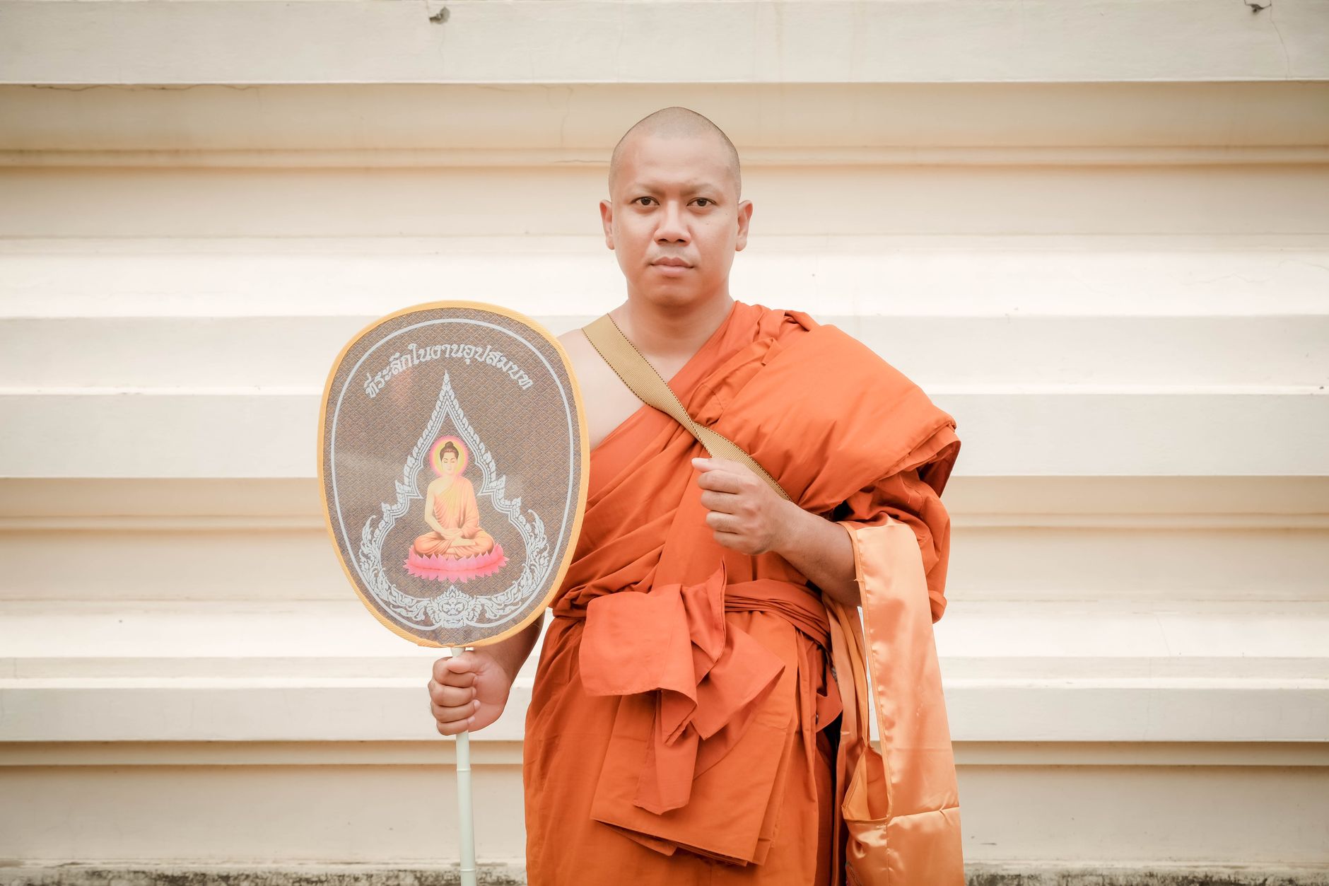 A Buddhist monk in traditional orange robes holding a ceremonial fan.