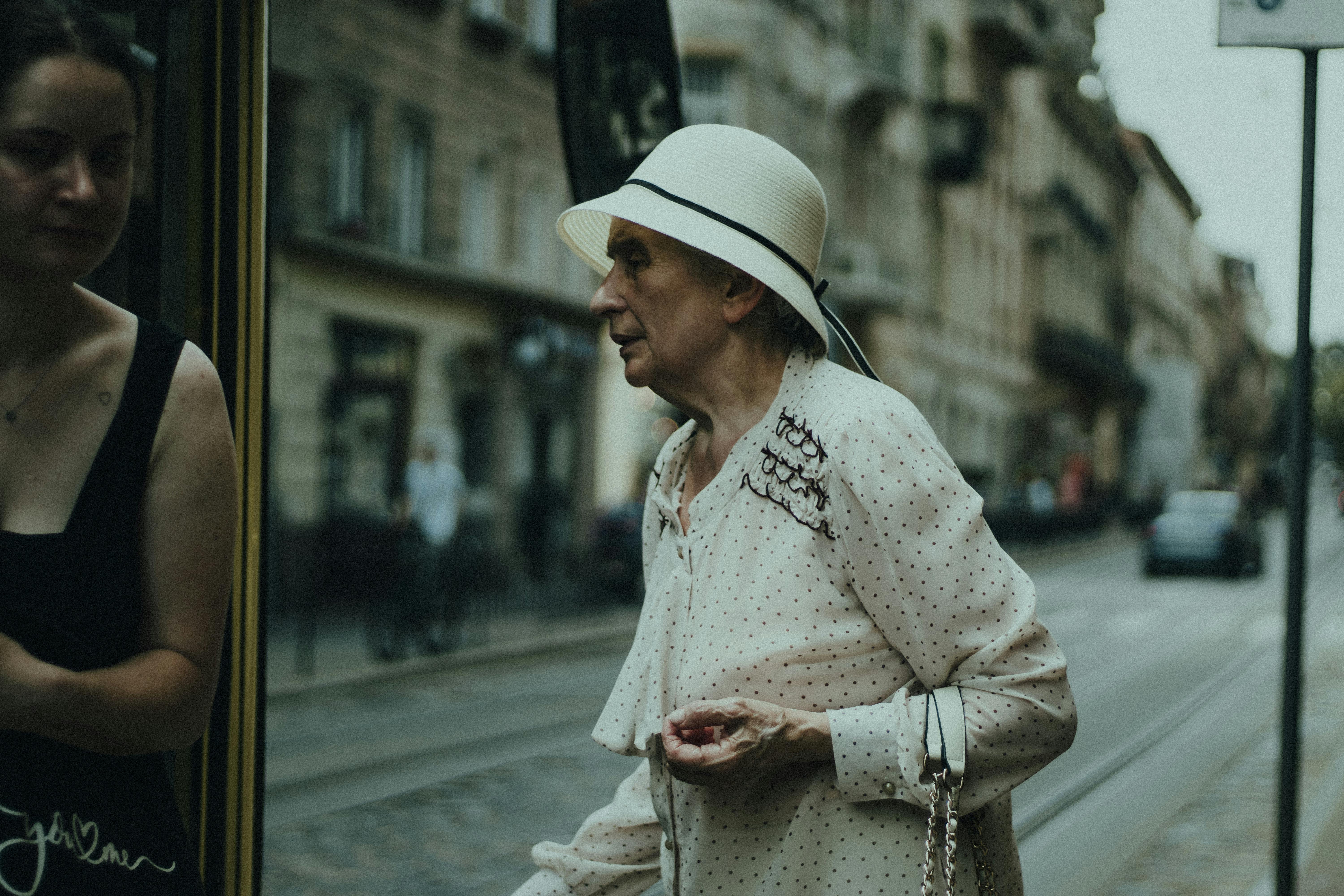 Elderly woman in fashionable attire walking on a city street, reflecting in a window.