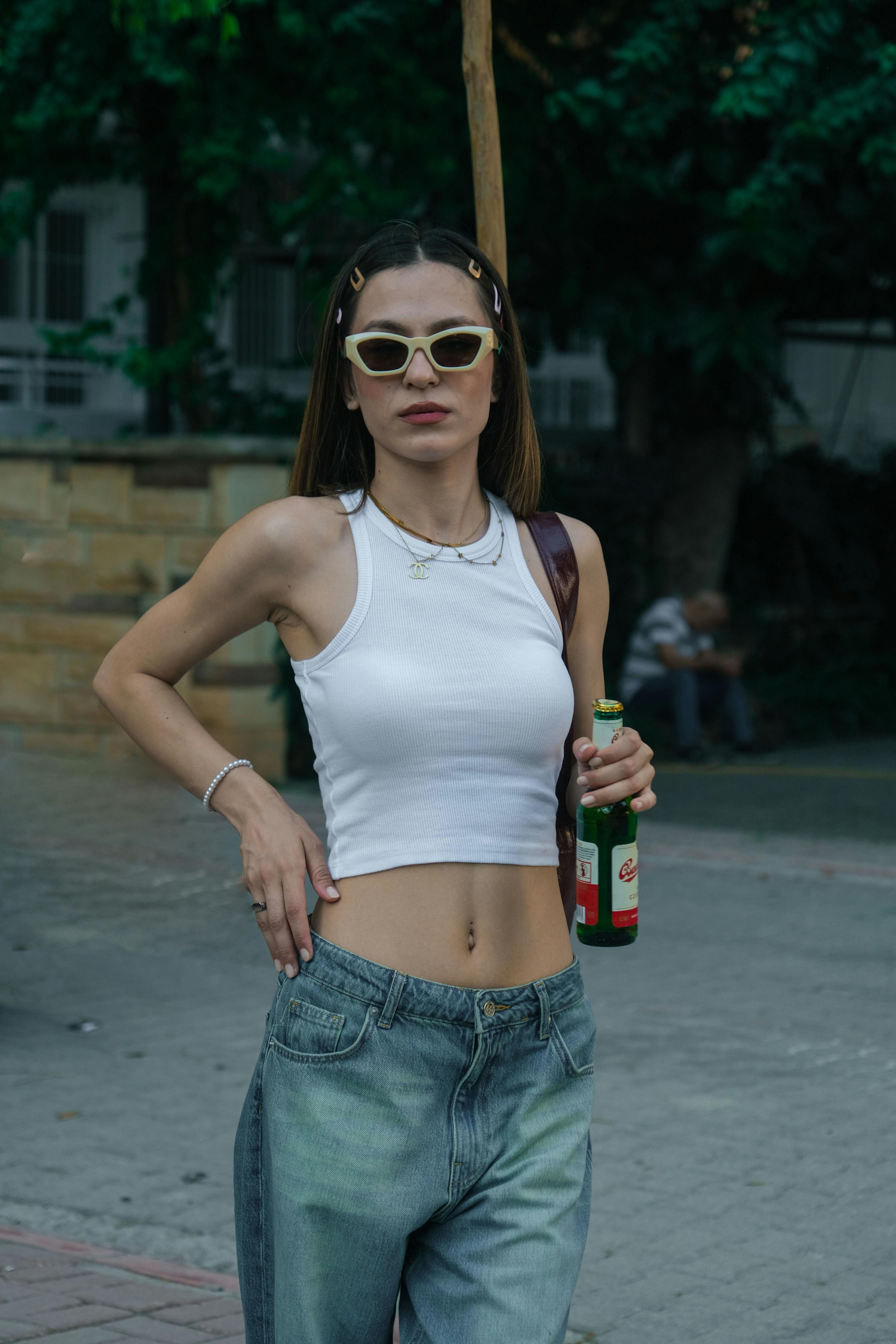Young woman in trendy attire holding a beverage bottle on a city sidewalk.