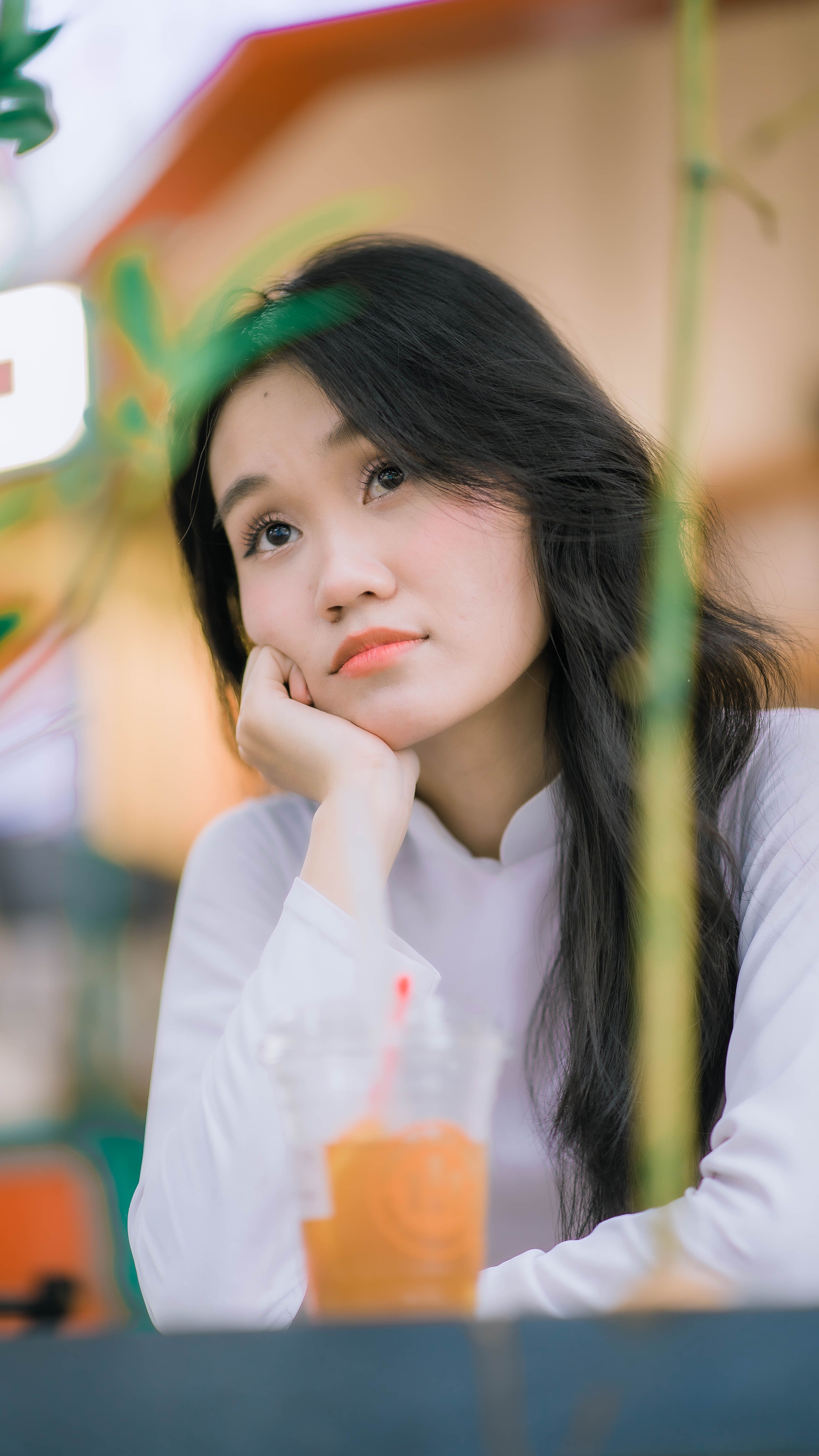 Thoughtful portrait of a young Asian woman sitting indoors with a drink, daydreaming.