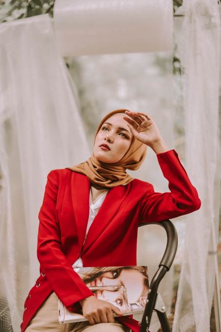 Elegant woman in a red blazer and headscarf, posing outdoors with a thoughtful expression.