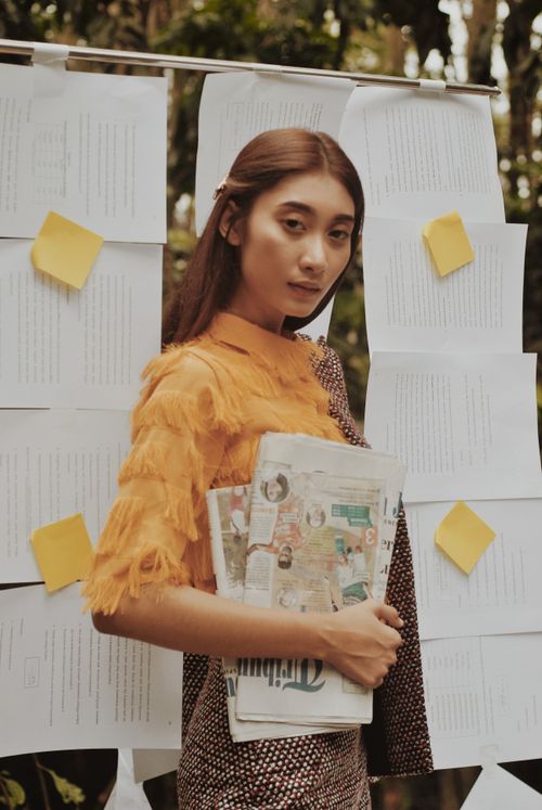 Free Stylish young woman poses with a newspaper in an outdoor setting with papers in the background. Stock Photo