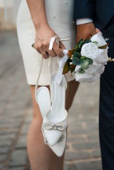 Close-up of bride holding bouquet and shoes alongside groom on wedding day.