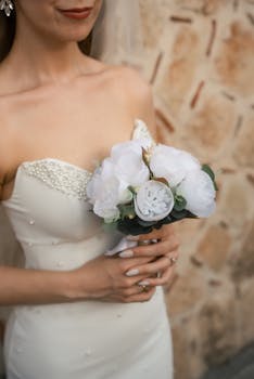 Bride in wedding dress holding a white bouquet against a rustic background.