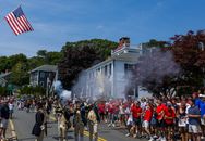 Crowded parade with reenactors in uniform firing rifles, surrounded by festive people waving flags on a sunny day.