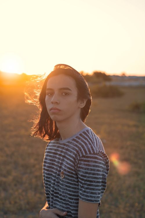Free Side view of peaceful young person with dark hair in casual clothes chilling on dry grassy meadow during sunset and looking at camera Stock Photo