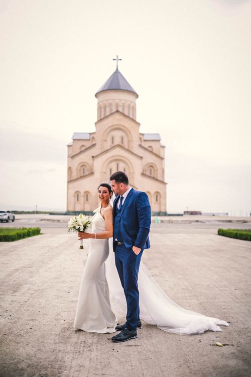 Free Newlyweds in elegant attire pose outside a grand church, embracing romance and style. Stock Photo
