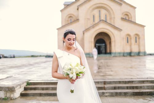 Free A beautiful bride in a white dress holding a bouquet in front of a church. Stock Photo