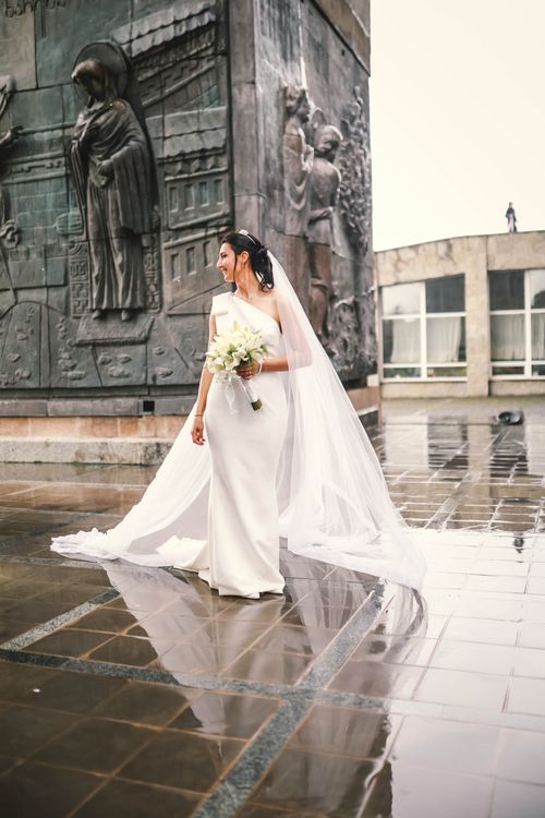 Free Bride in white dress and veil holding bouquet near an urban statue. Stock Photo