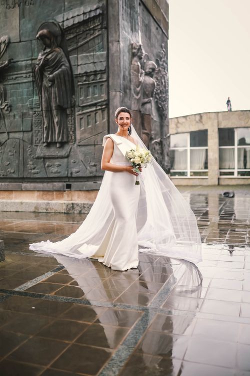Free Bride in a white dress with a veil holding a bouquet, standing near a monument on a rainy day. Stock Photo