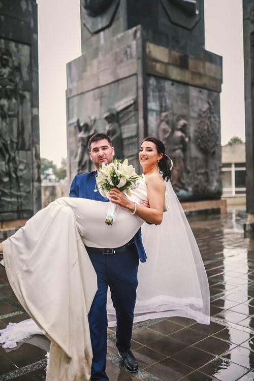 Free Bride in groom's arms with a city monument backdrop, symbolizing love and celebration. Stock Photo