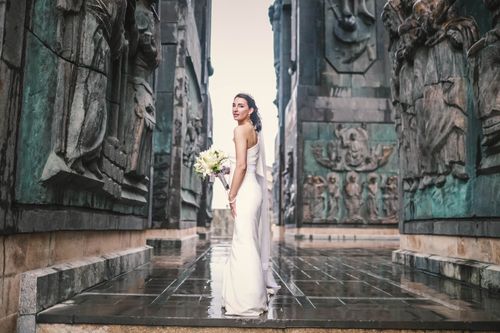 Free Bride in a wedding dress stands elegantly with a bouquet, framed by monumental sculptures. Stock Photo