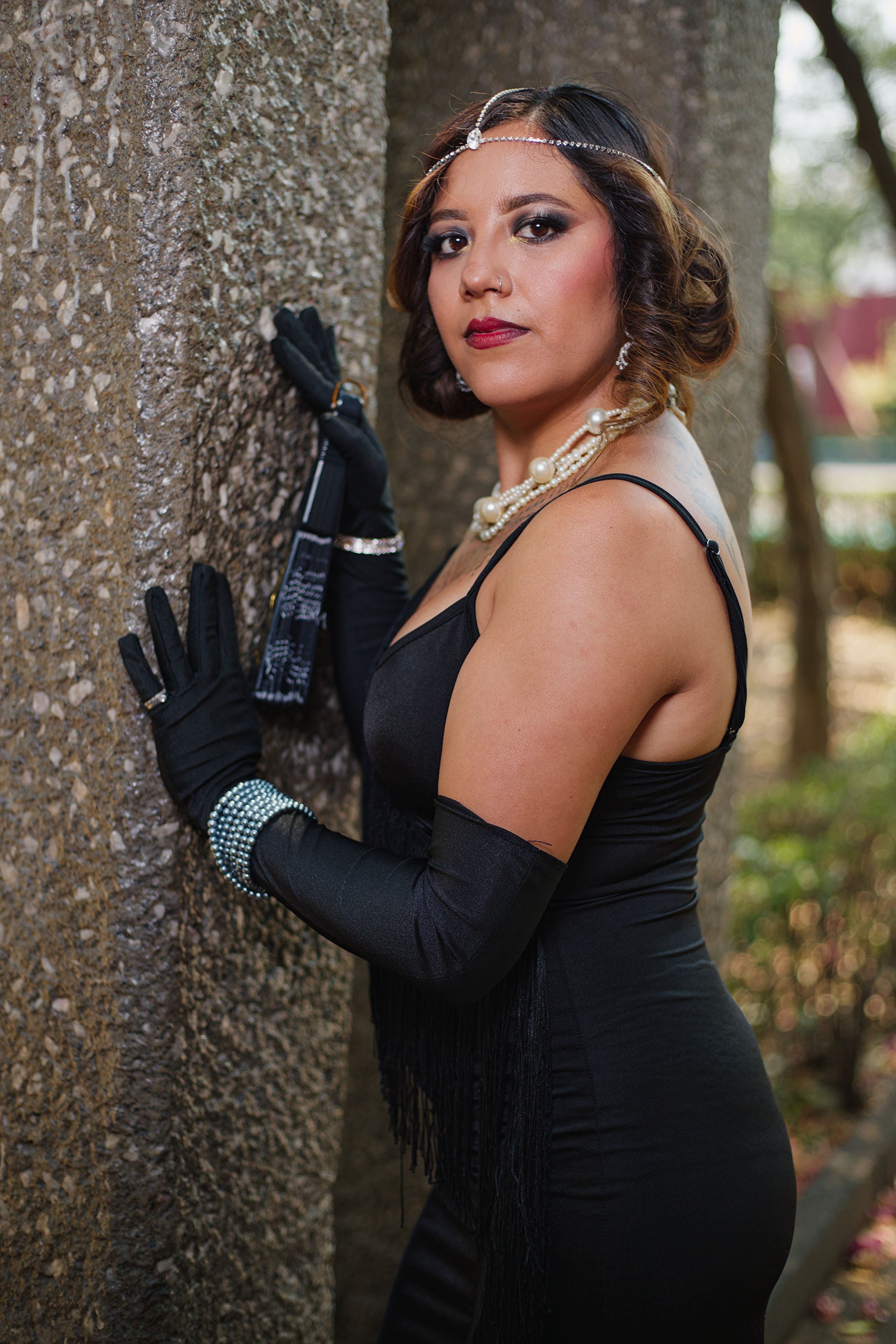 Woman in vintage 1920s fashion with pearls and headdress posing outdoors against a wall.