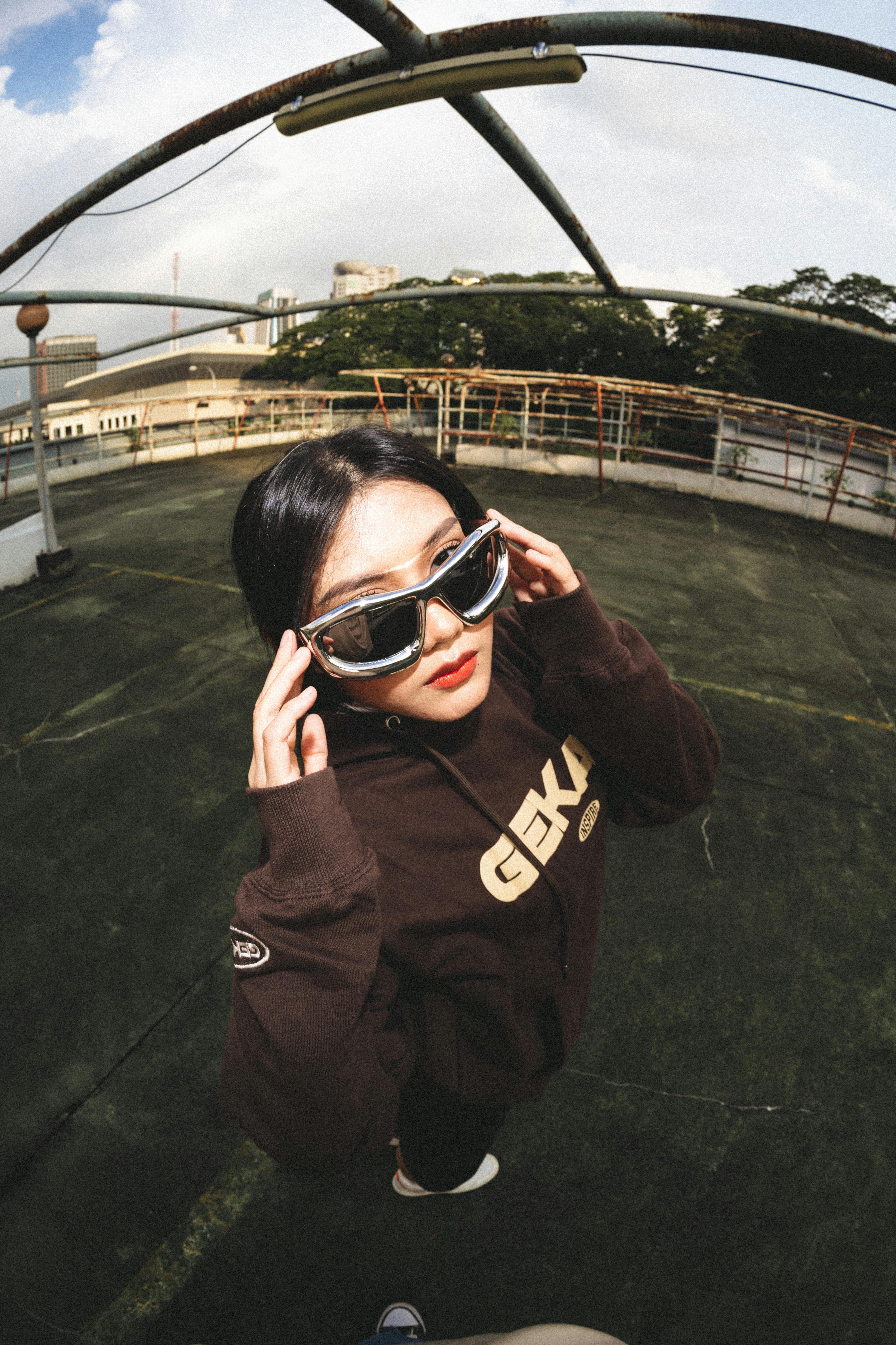 Young woman in stylish sunglasses poses on rooftop, fisheye lens effect.