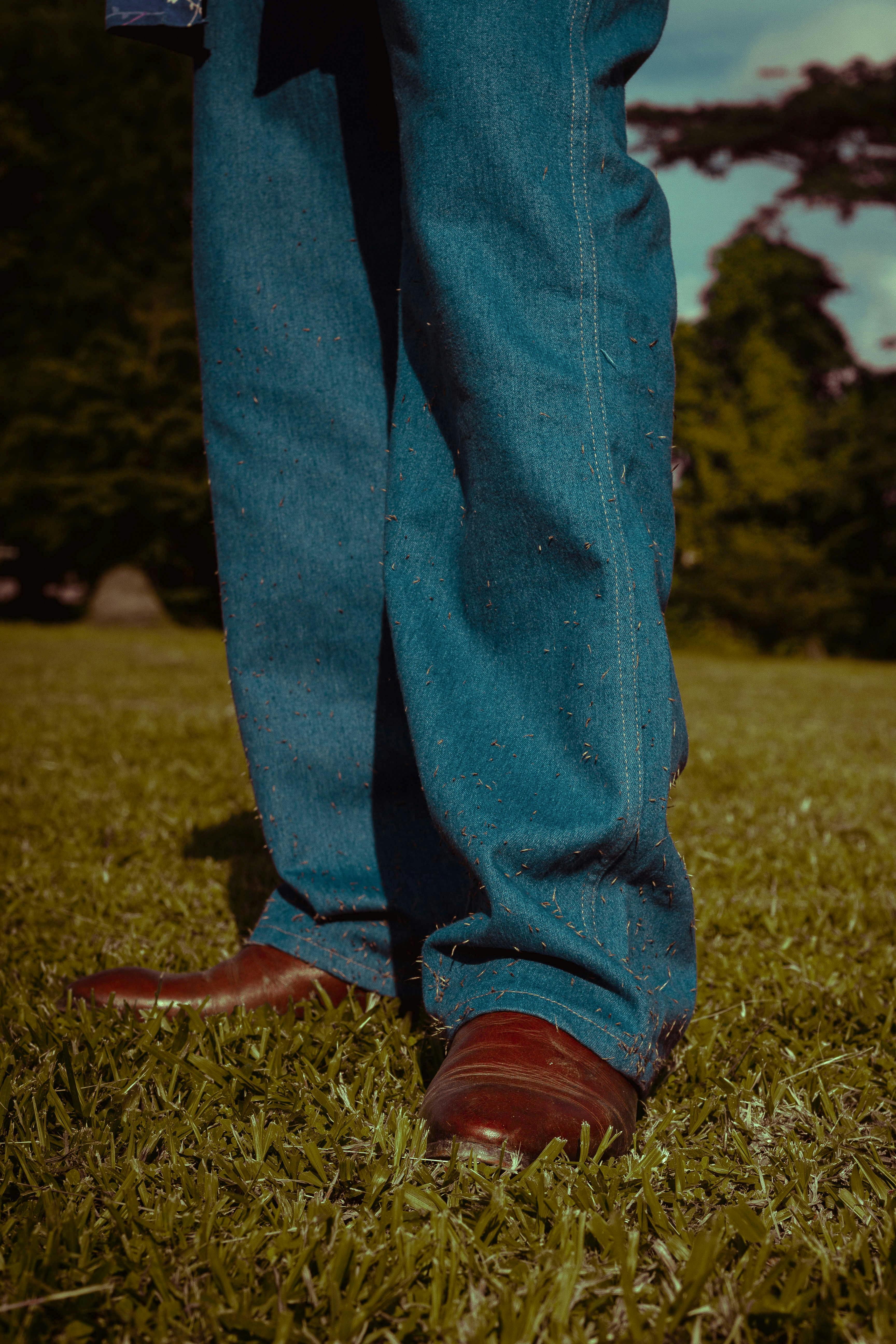 Close-up of a man in denim and brown shoes standing on grass outdoors.