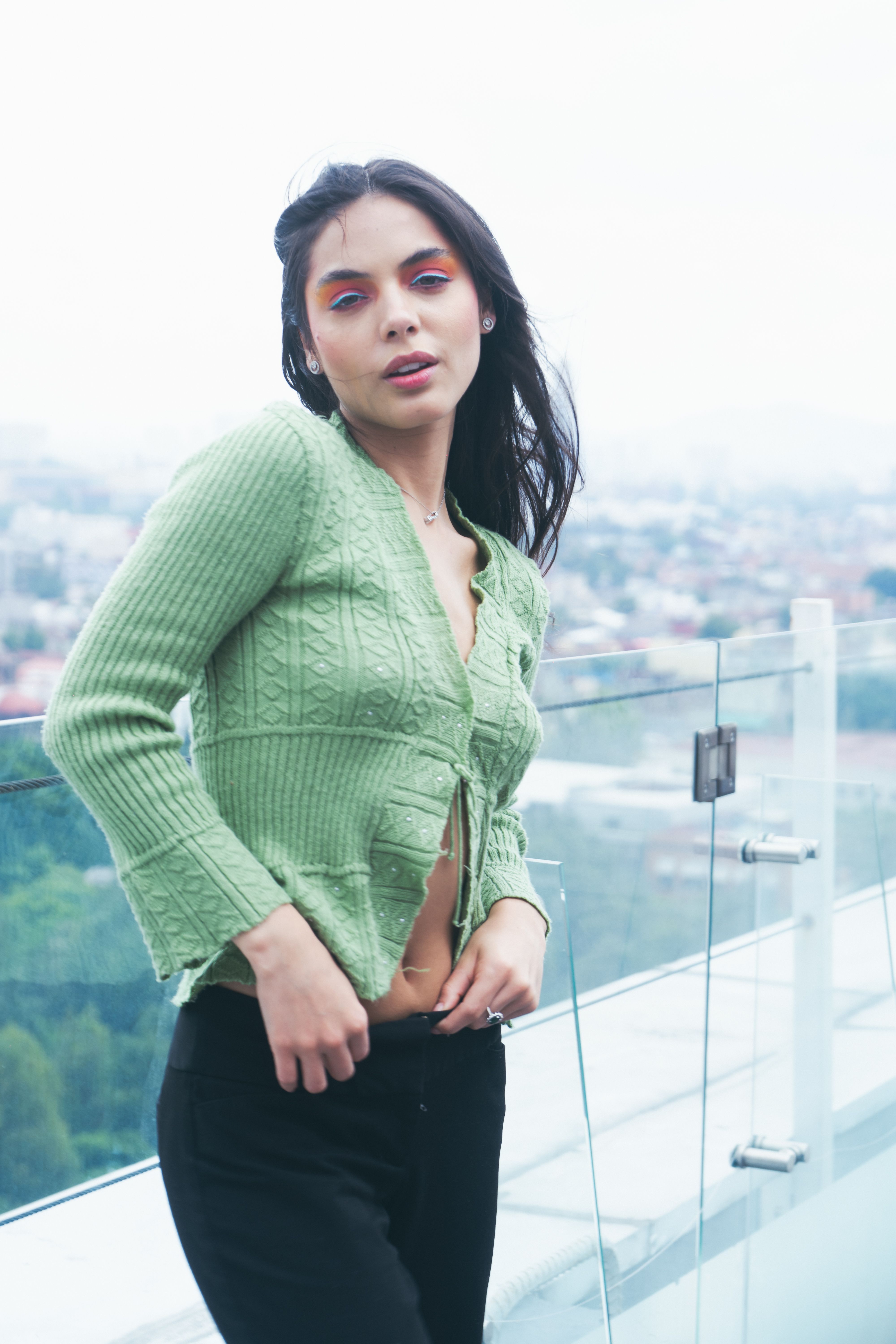 Portrait of a fashionable woman in a green sweater, posing elegantly on a rooftop with a city view.