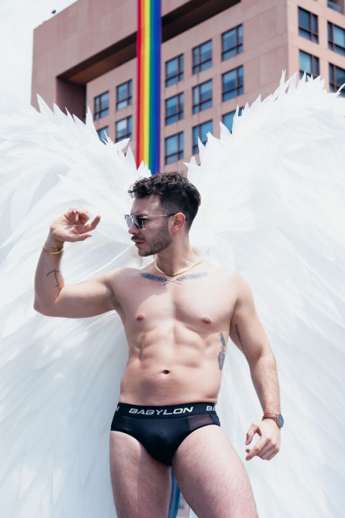 Free Model with angel wings poses confidently in an urban setting with a rainbow banner. Stock Photo