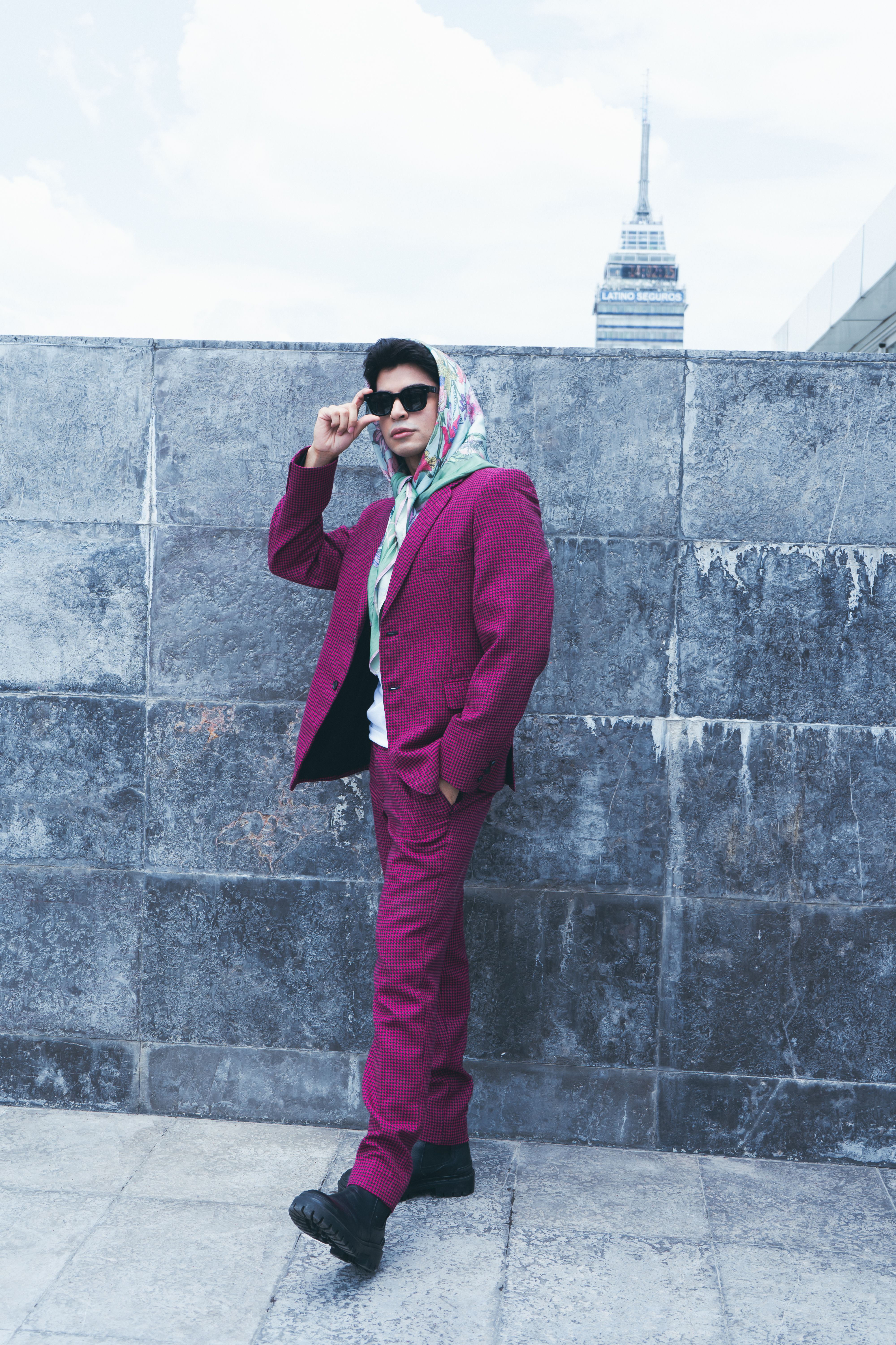 Fashionable man in pink suit poses with sunglasses against a stone wall in Mexico City.