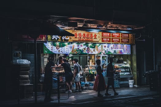 A bustling urban street food stall illuminated at night with passersby enjoying snacks.