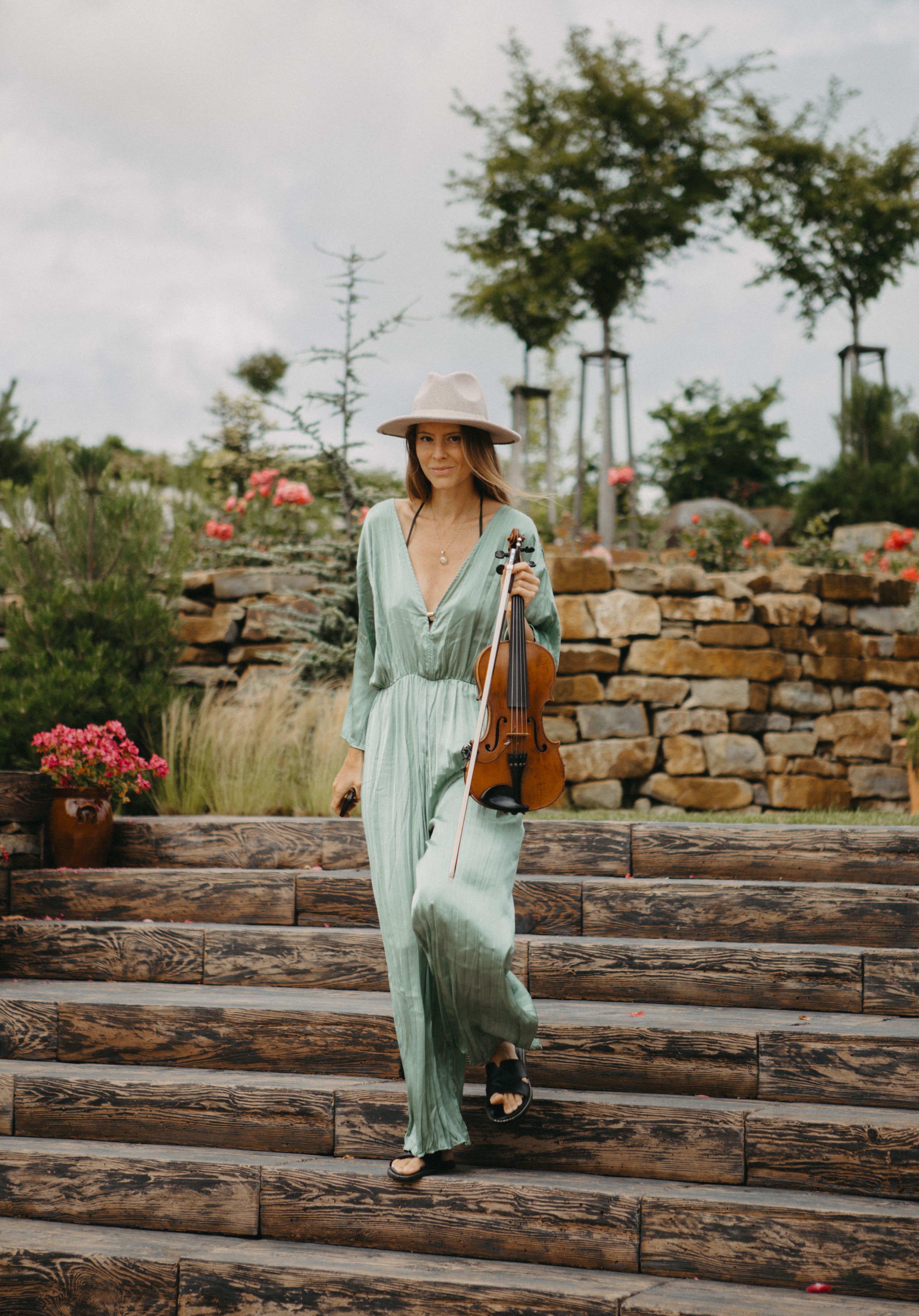 A woman in a green dress walking outdoors, holding a violin, in a lush garden with stone steps.