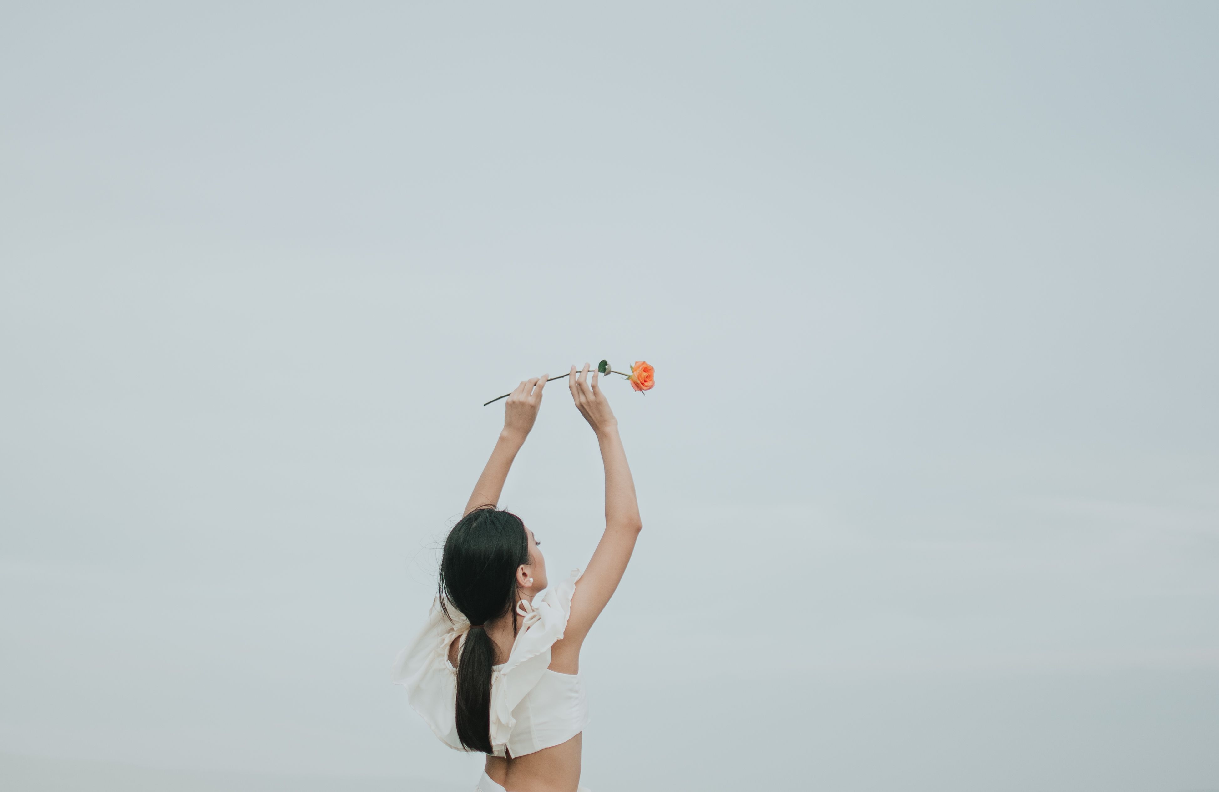 一名女子高举一朵橙色玫瑰，在柔和天空的映衬下，尽显优雅与自由