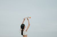 一名女子高举一朵橙色玫瑰，在柔和天空的映衬下，尽显优雅与自由