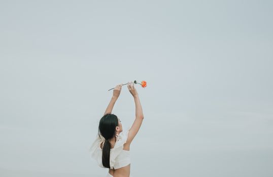 一名女子高举一朵橙色玫瑰，在柔和天空的映衬下，尽显优雅与自由