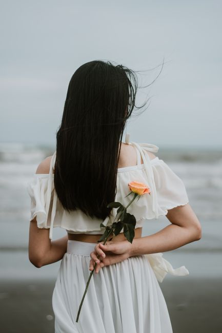 A woman with long hair holds a rose on a serene beach, embodying elegance and tranquility.