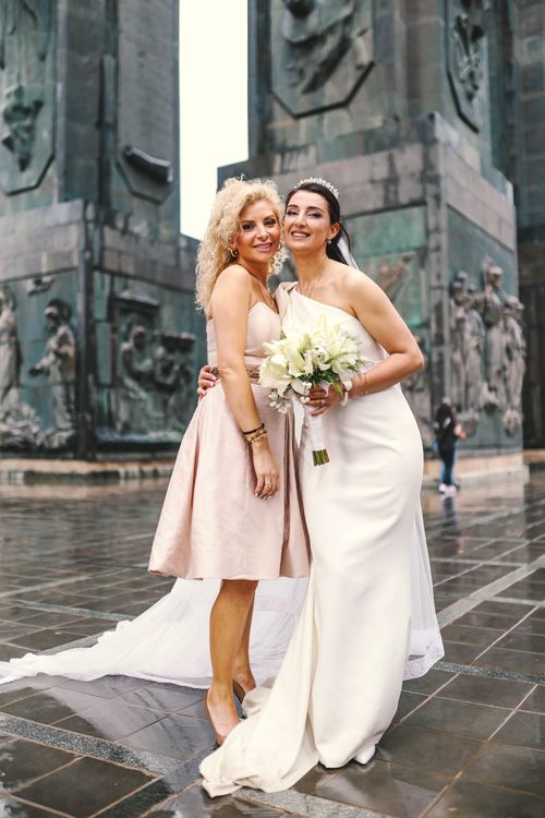 Free Bride and bridesmaid in elegant dresses posing with smiles in an outdoor stone architecture setting. Stock Photo