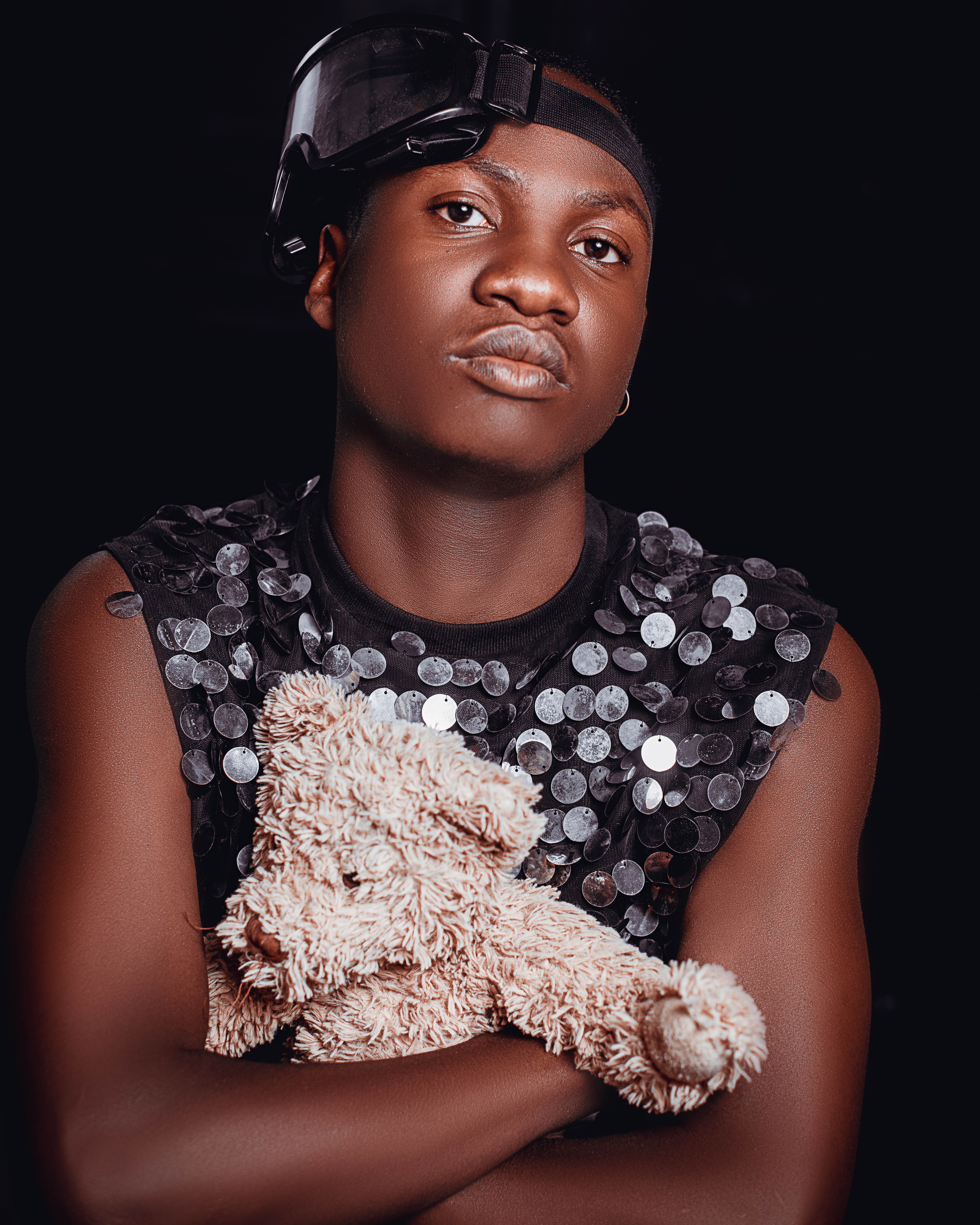 Fashionable portrait of an adult African man in sequined top holding a teddy bear against a dark backdrop.