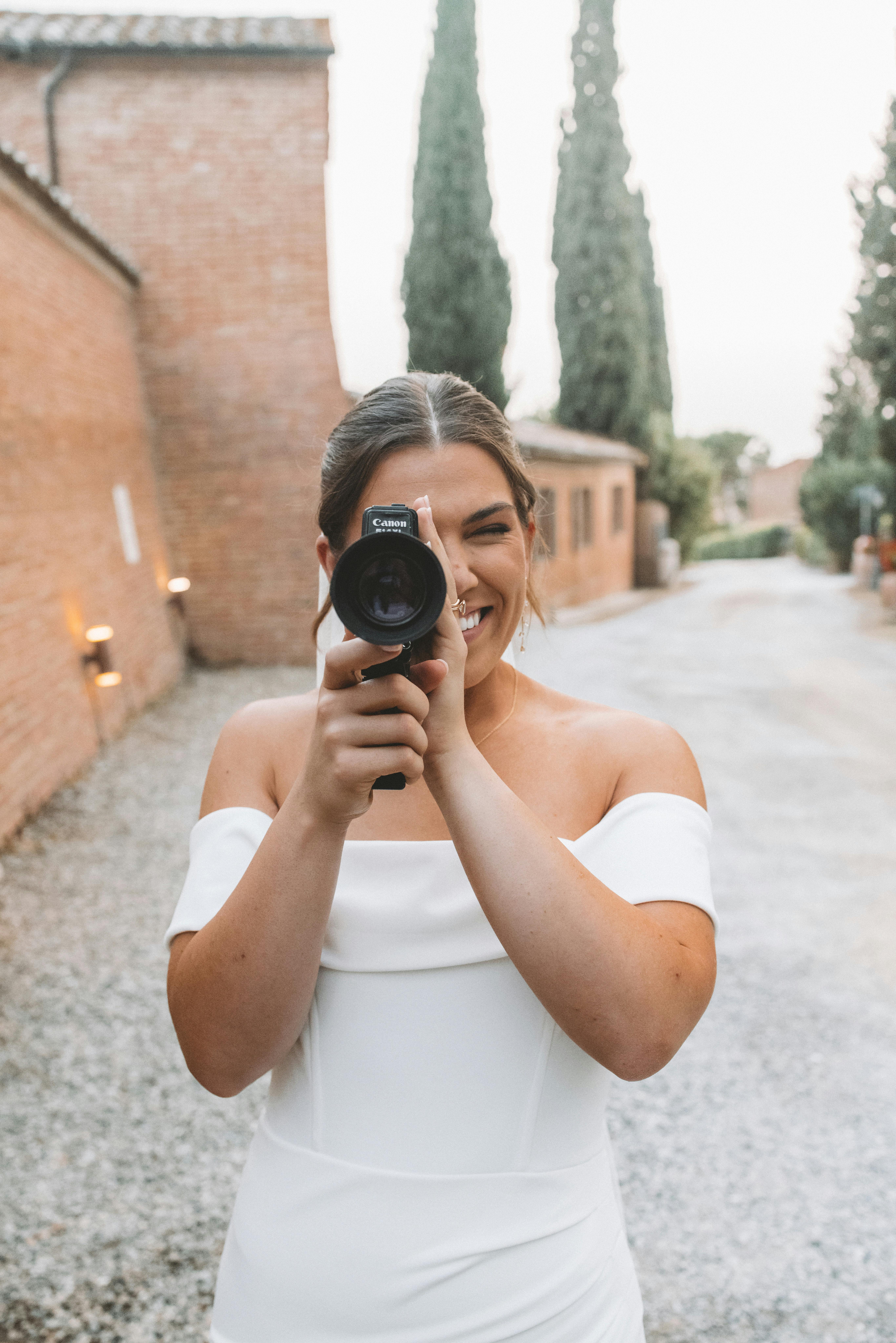 A smiling bride in a white dress playfully holds a camera in a charming rustic setting.