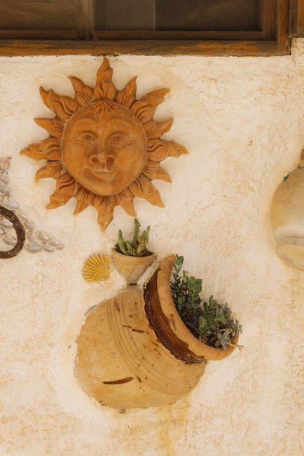 Close-up of a sun face sculpture with clay pot and plants on a rustic wall.